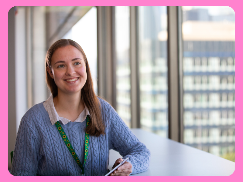 A woman wearing a blue cable-knit sweater over a collared shirt sits at a table in front of large windows showing a cityscape. She has a green lanyard around her neck and is holding a tablet.