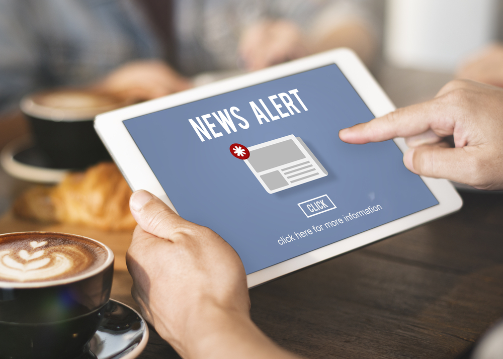 A person holding a tablet showing a 'NEWS ALERT' screen with a newspaper icon and the word 'CLICK' beneath it. The bottom of the screen reads 'click here for more information.' In the background, blurred figures and a coffee cup are visible on a table.