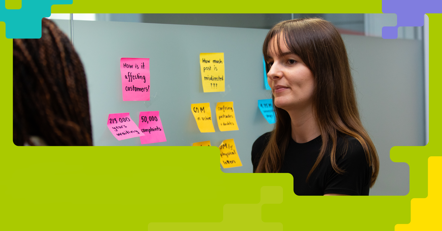 Two people stand in front of a glass wall covered with colourful sticky notes containing handwritten questions and statements, such as 'How is it affecting customers?' and 'How much post is misdirected???'. The scene suggests a collaborative brainstorming or planning session in a work environment.