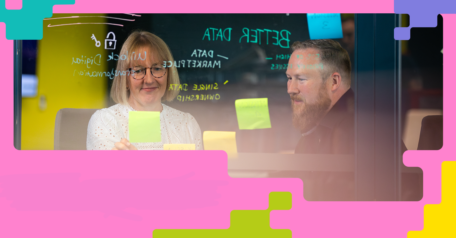 A woman and a man are sitting at a table in an office setting, discussing or collaborating in front of a glass wall covered with sticky notes and phrases like 'BETTER DATA' and 'Unlock Digital Transformation.' The image is framed with colourful geometric shapes, suggesting a creative and innovative workspace.