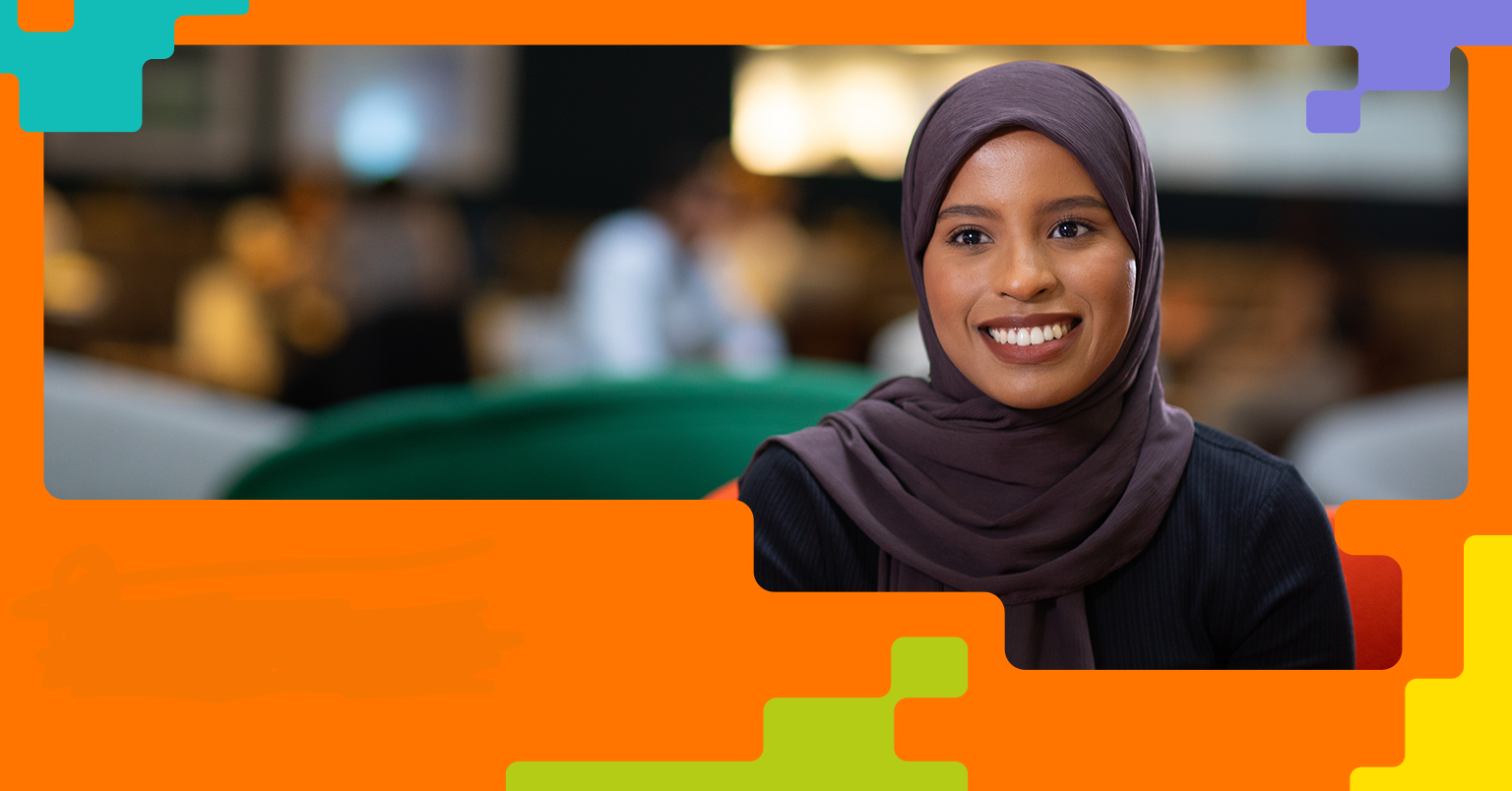 A woman wearing a headscarf and dark clothing smiles in an indoor setting with a blurred background. The image is framed by colourful geometric shapes, highlighting the individual in a professional or public environment.