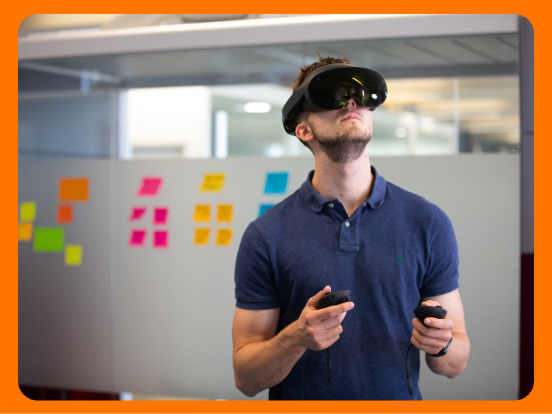 A man in an office setting wearing a virtual reality headset and holding controllers. The background features colourful sticky notes on the wall and various office furniture.