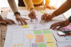 Group of people collaborating around a table covered with documents, charts, and sticky notes, with several hands visible indicating active discussion and teamwork.