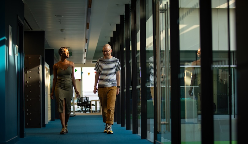 A man and a woman walking down a modern office corridor with glass walls on one side and lockers on the other. The hallway has blue carpeting, and there is natural light coming from large windows. In the background, an office area with chairs and desks is visible.
