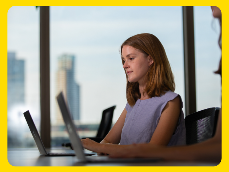 A woman seated at a desk working on a laptop in a modern office setting. The background shows large windows with an urban cityscape and tall buildings visible outside. The individual is wearing a sleeveless top and is positioned in front of another laptop, suggesting a collaborative or professional environment.