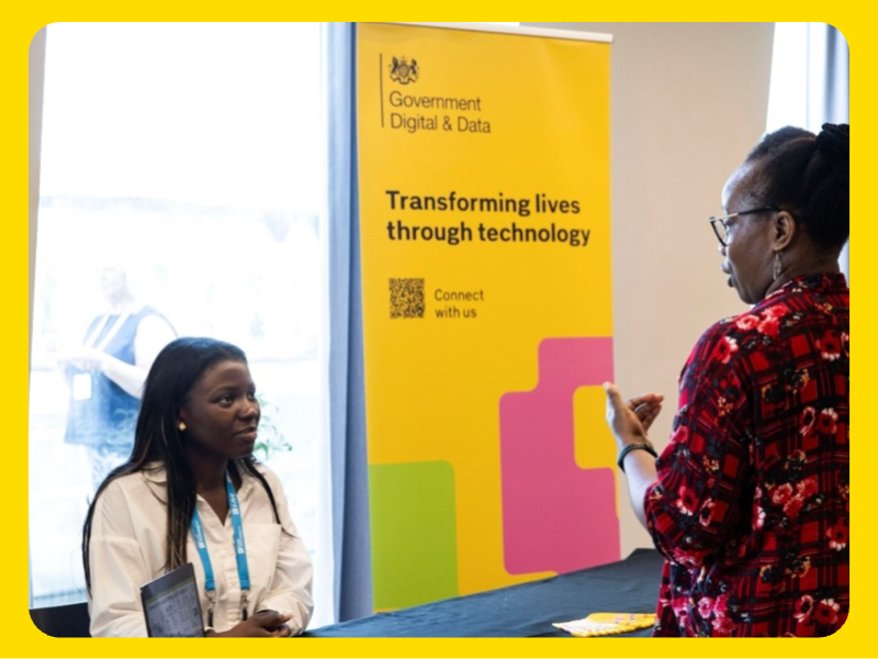 Two women are engaged in conversation at an event booth. Behind them is a bright yellow banner with the UK Government Digital & Data logo and the text: “Transforming lives through technology” along with “Connect with us.” The banner features coluorful geometric shapes in pink and green. A table in front of the banner holds informational materials. The setting appears to be a professional networking or recruitment event.