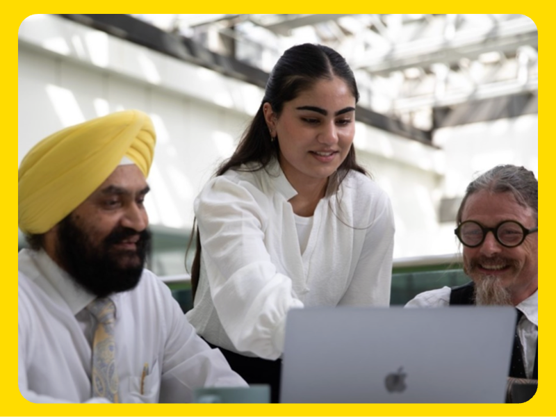 Two men and one woman are gathered around a laptop in a bright, modern indoor setting with a glass ceiling structure visible above. The woman is standing and pointing at the laptop screen, while the men are seated and looking toward the device. The group appears to be engaged in a collaborative discussion or review of work. The scene conveys teamwork and professional interaction.