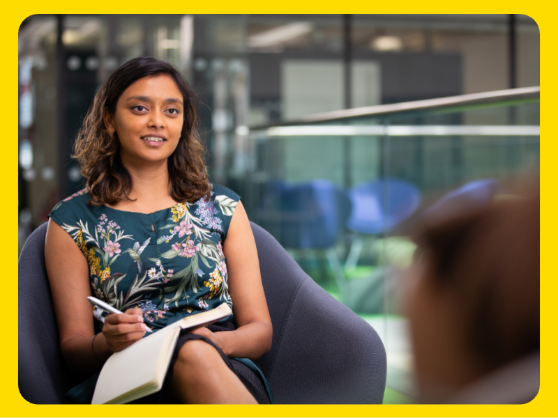 A woman is seated in a modern indoor setting, holding a pen and an open notebook, appearing to take notes or prepare for a discussion. The woman is wearing a dark floral-patterned top and is sitting in a curved chair. The background shows a glass railing and blue chairs, suggesting an office or meeting space.