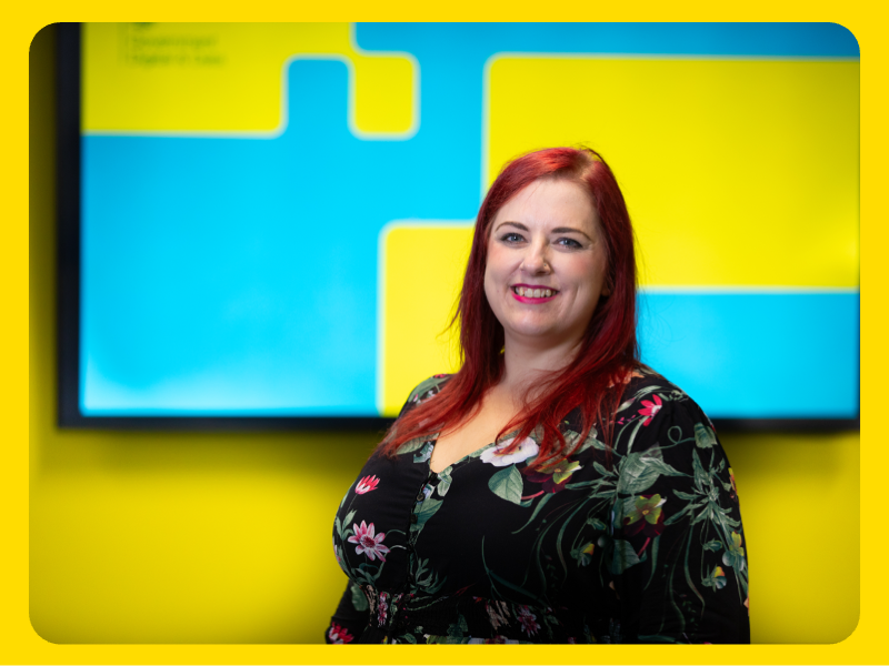 A woman is standing indoors in front of a bright yellow and blue background featuring geometric shapes. The background appears to be part of a display or presentation screen. The woman is wearing a dark floral-patterned top and has long red hair. The setting suggests a professional or event environment.