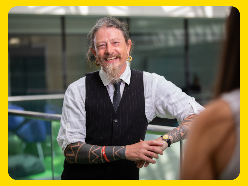 A man is wearing a white shirt with rolled-up sleeves, a black pinstripe vest, and a black tie is leaning casually against a railing in a modern indoor setting. The man has visible tattoos on both forearms, a wristwatch, and a red wristband. The background includes glass panels and blurred seating, suggesting an office or collaborative space.