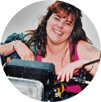 A woman wearing a pink top and denim jacket, sitting on a big motorbike with a large headlight and visible handlebars.