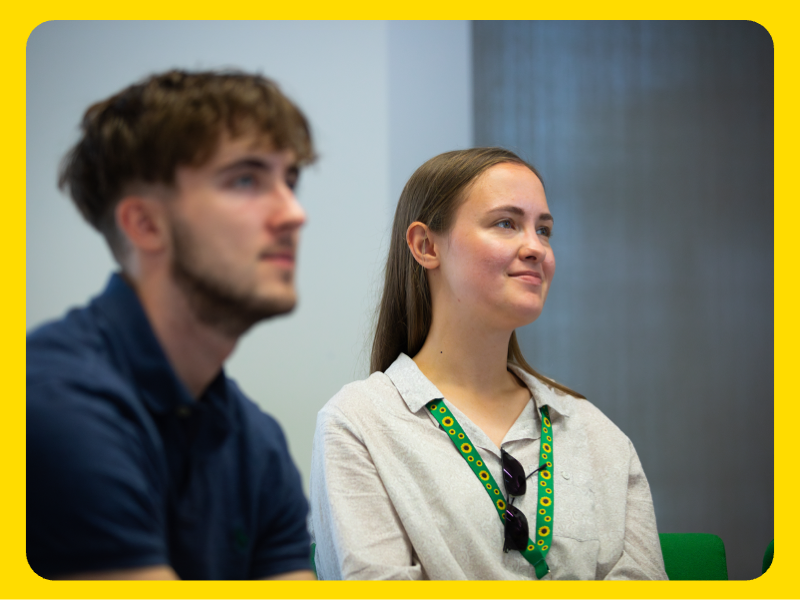 A man and a woman are seated indoors, appearing to listen attentively during a discussion or presentation. The woman is wearing a light-coloured shirt with a green lanyard and sunglasses hanging from it, while the man is dressed in a dark shirt. The background shows a neutral wall and part of a green chair, suggesting a professional or meeting environment.