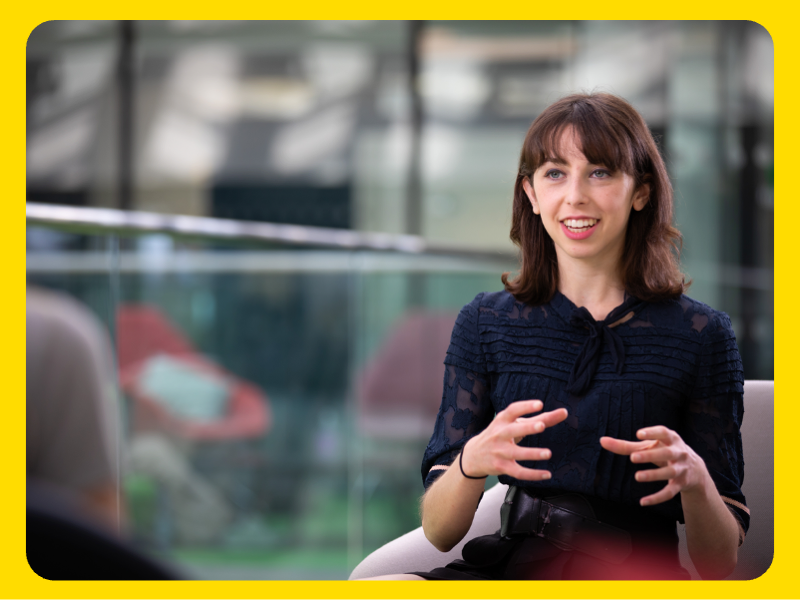 A woman is seated indoors, wearing a dark textured top, gesturing with both hands while speaking. The background shows a modern setting with glass panels and blurred furniture.