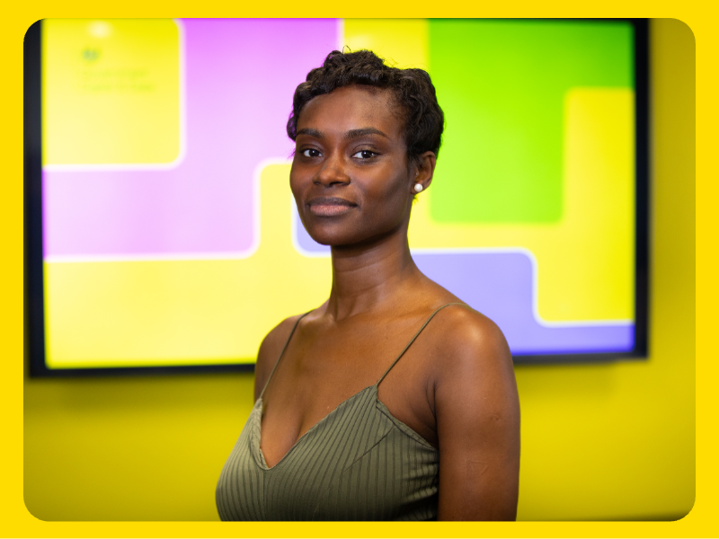 A woman is standing indoors in front of a bright display screen featuring a coluorful geometric design in yellow, green, and purple. The woman is wearing a sleeveless, olive-green top with thin straps. The setting appears to be a professional or event environment.