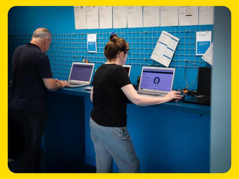 A man and a woman are standing at a bright blue counter, each working on a laptop. The counter has multiple laptops connected with cables, and a grid panel on the wall behind it holds various documents and papers, some pinned and others hanging. The setting appears to be a resource or help desk area, possibly for technical support or training.