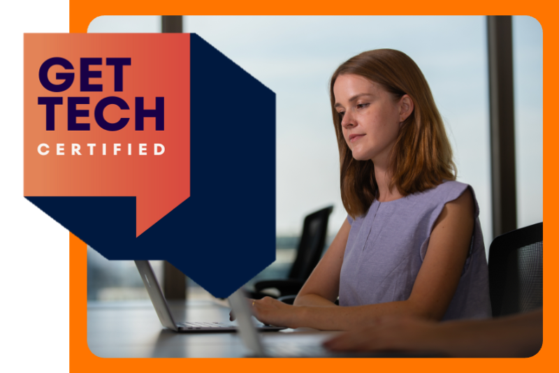 A woman seated at a desk working on a laptop in a modern office setting. The background shows large windows with an urban cityscape and tall buildings visible outside. The individual is wearing a sleeveless top and is positioned in front of another laptop, suggesting a collaborative or professional environment.