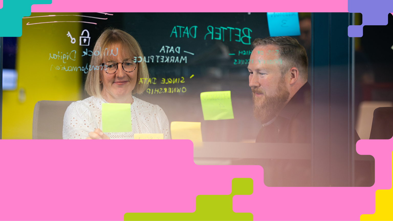 A woman and a man are sitting at a table in an office setting, discussing or collaborating in front of a glass wall covered with sticky notes and phrases like 'BETTER DATA' and 'Unlock Digital Transformation.' The image is framed with colourful geometric shapes, suggesting a creative and innovative workspace.