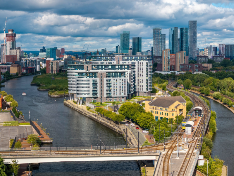 A wide aerial view of a modern urban waterfront area. A river curves through the scene with several bridges crossing it. In the centre is a cluster of contemporary mid‑rise apartment buildings with grey and white facades. To the right, elevated rail tracks carry a light‑rail train toward a small station surrounded by trees. The foreground includes roads, bridges, and greenery, while the background shows a dense skyline of tall glass skyscrapers under a dramatic cloudy sky.