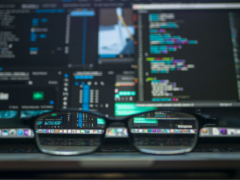 A pair of glasses rests in front of a laptop screen displaying lines of colourful programming code and system dashboards, with the code reflected sharply in the lenses.