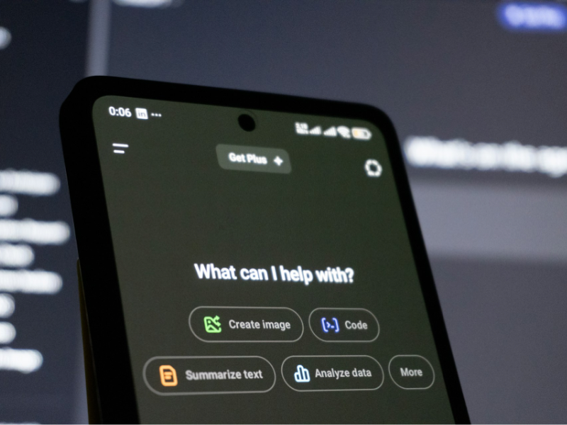 A smartphone screen displays an AI assistant interface with options such as “Create image,” “Code,” “Summarize text,” and “Analyze data,” set against a blurred background of a computer display.