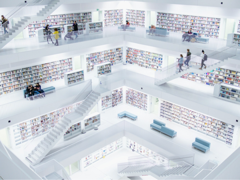 A large, bright, multi‑level modern library with white interiors, open staircases, and rows of bookshelves lining each floor, with a few people walking or reading throughout the space.