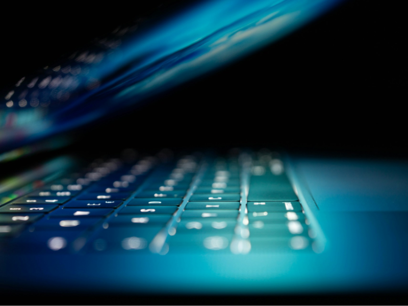 A close-up, low‑angle view of a laptop keyboard glowing in blue light, with the screen partially closed in a dark environment.