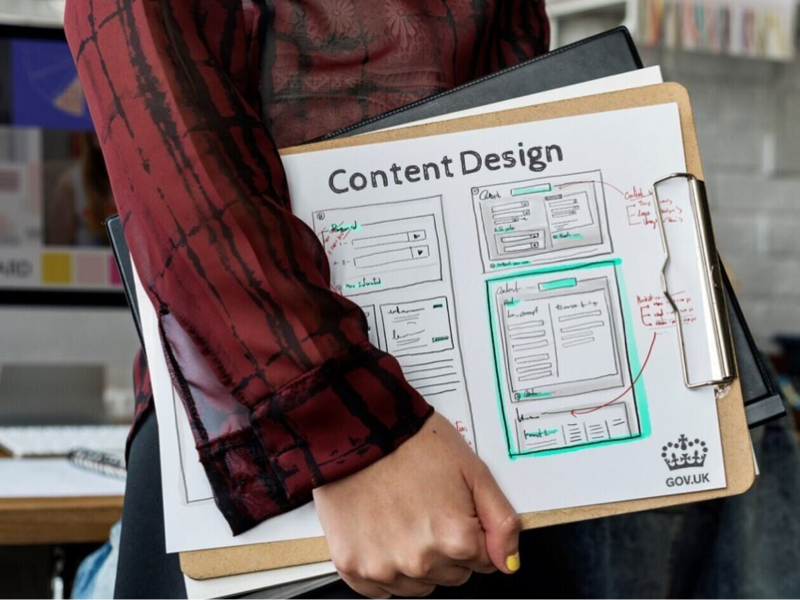 A person holding a clipboard with a printed “Content Design” mock‑up that includes hand‑drawn website wireframes and annotations, alongside a GOV.UK logo.