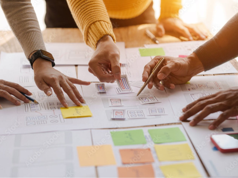 Several people collaborating over a table covered with sketched website wireframes and colourful sticky notes, pointing to and arranging paper interface elements as part of a design or user‑research workshop.