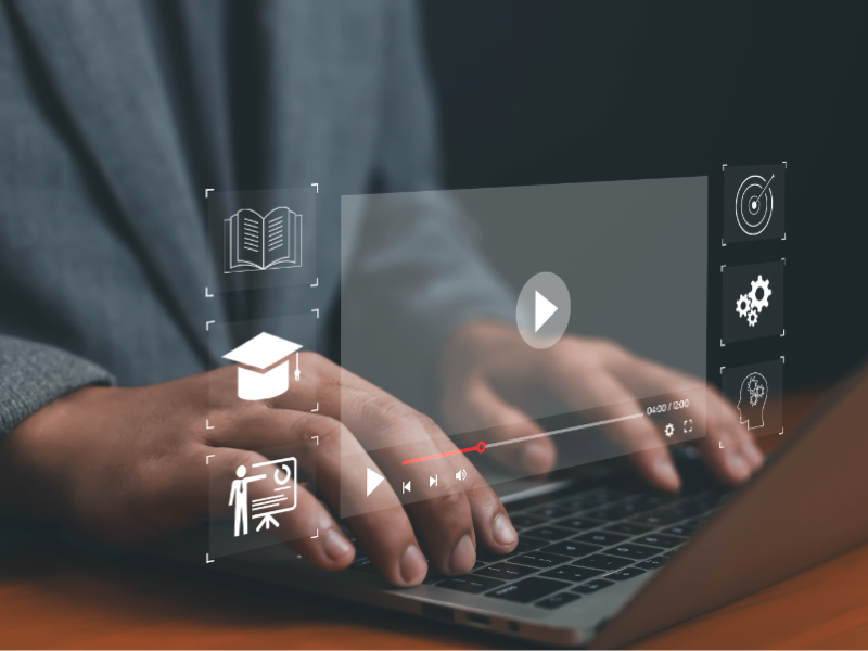 Close‑up of a person’s hands typing on a laptop keyboard, with semi‑transparent digital interface icons overlaid. The icons include an open book, a graduation cap, a presenter at a board, a play button on a video player timeline, a target, gears, and a lightbulb. The scene suggests online learning or digital training.